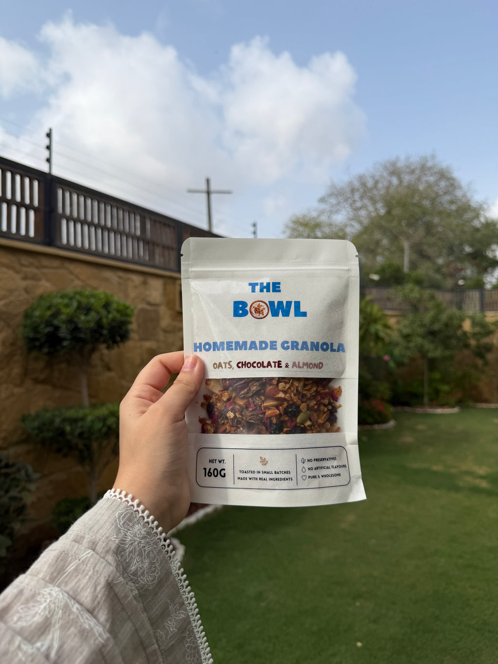 Hand holding a package of 'The Bowl' homemade granola with a garden and sky in the background 